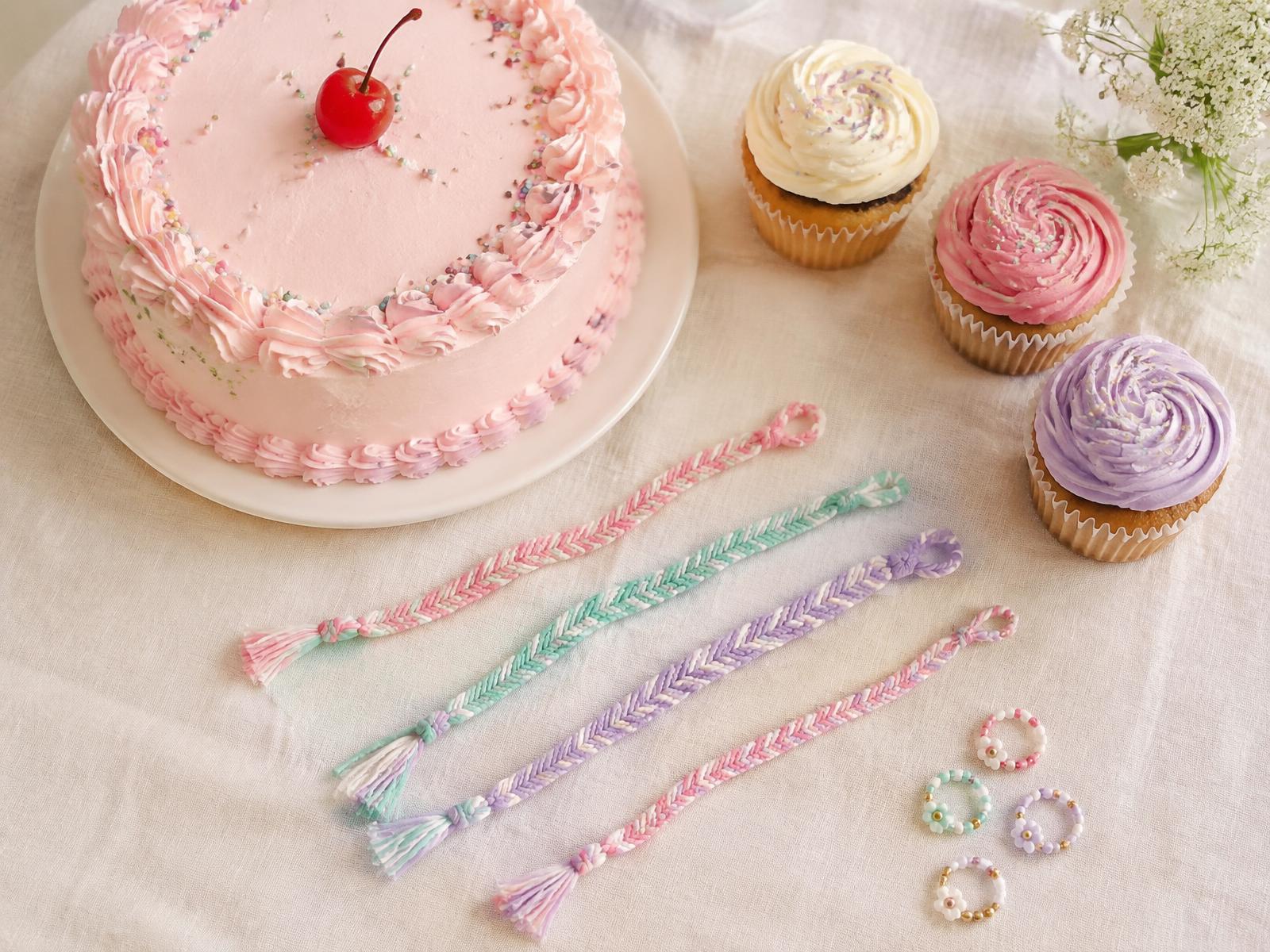 A pink celebration cake and frosted cupcakes beside handmade fishtail bracelets and beaded rings